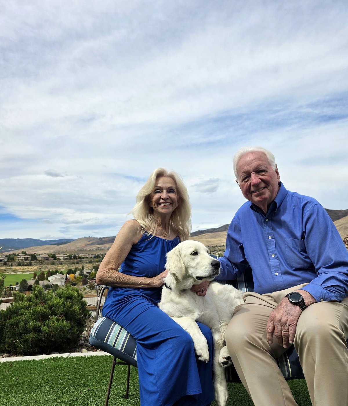 Bill and Mary O’Donnell smiling on a garden bench with Maggie, their cream golden retriever puppy, under a big sky, with Somersett’s fairway and golden hills behind them.